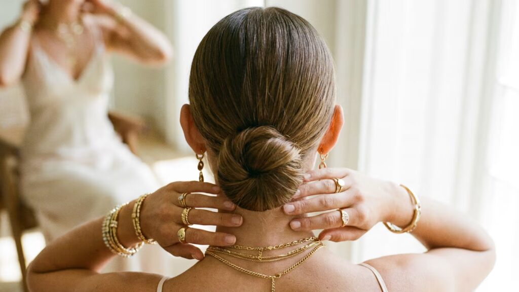 Oreilles dégagées, chignon bas et accumulation élégante de bijoux dorés, avec la robe claire en arrière-plan.